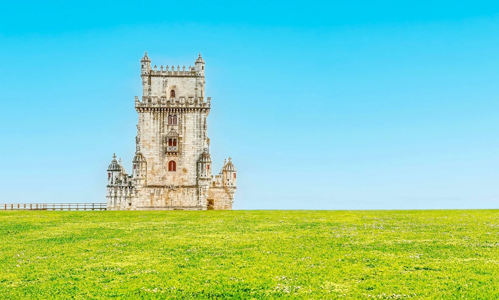 Belém Tower in pale limestone standing at the edge of a wide green lawn under a clear blue sky