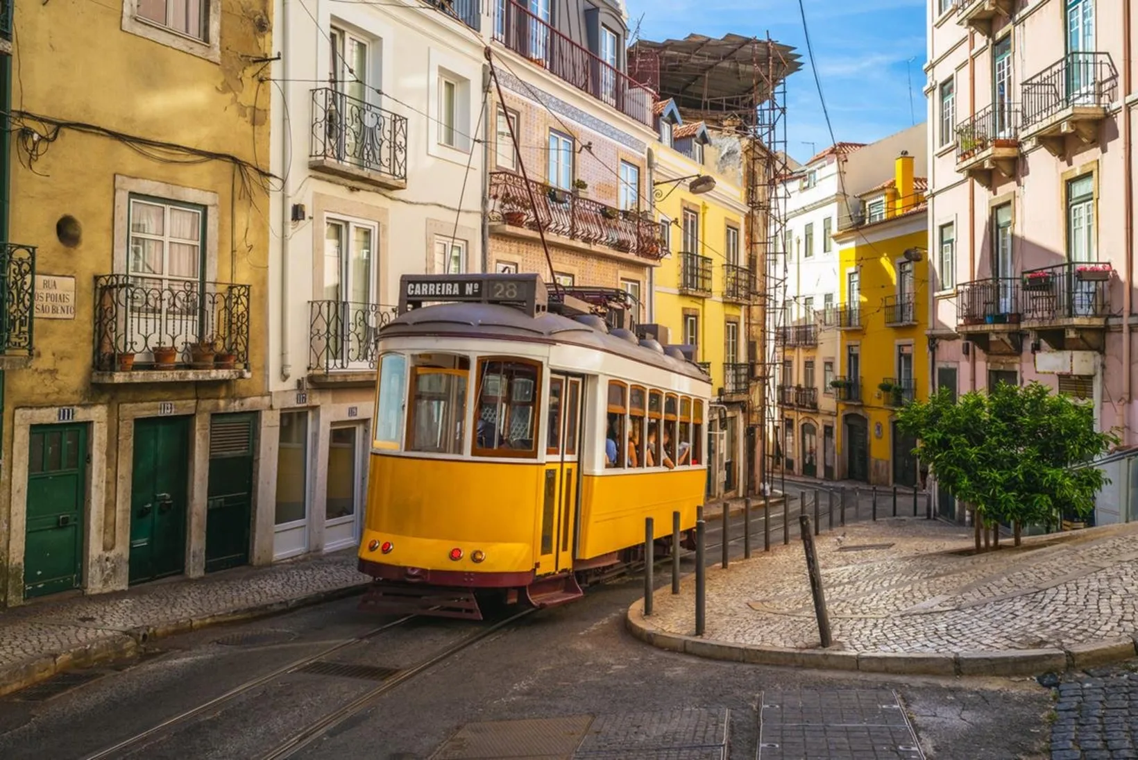 A yellow Tram 28 streetcar rounding a narrow cobbled corner between pastel tiled buildings in Alfama
